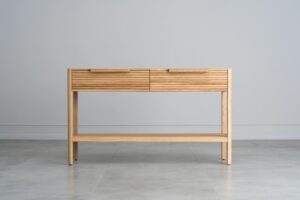 Tento odkládací stolek z masivního dubu kombinuje funkčnost s elegantním designem. Má dvě praktické zásuvky s charakteristickým drážkovaným čelem a minimalistickými úchyty, které dodávají moderní vzhled.